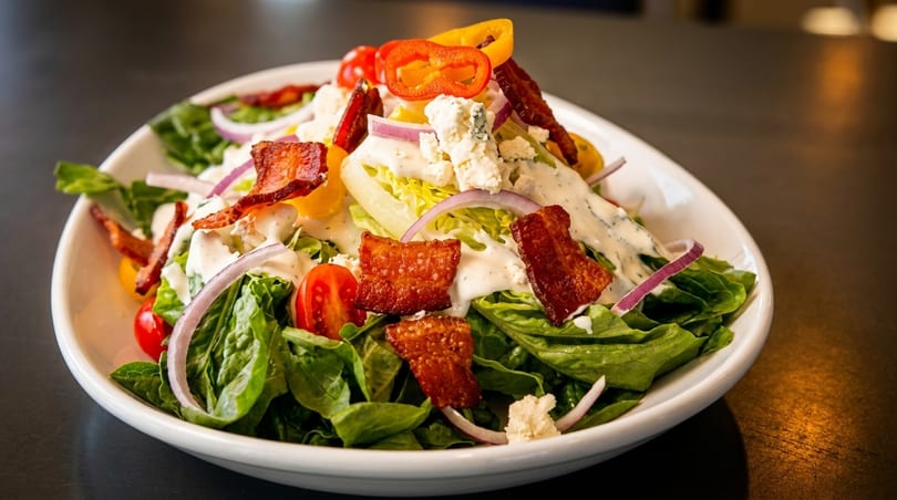 Fresh green salad with bacon, blue cheese, red onions, tomatoes, and bell peppers, drizzled with dressing.