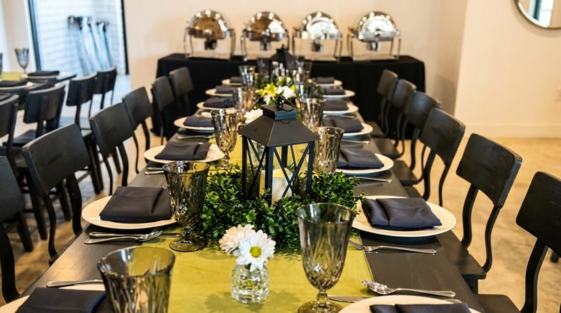 Long dining table set with dark chairs, green runner, black lantern, and buffet chafing dishes.