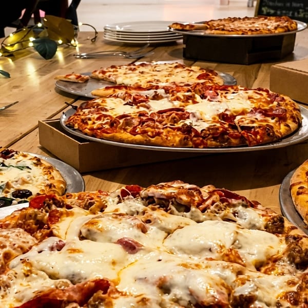 A wooden table laden with several large, hot pizzas, including pepperoni, white sauce, and meat lover's.