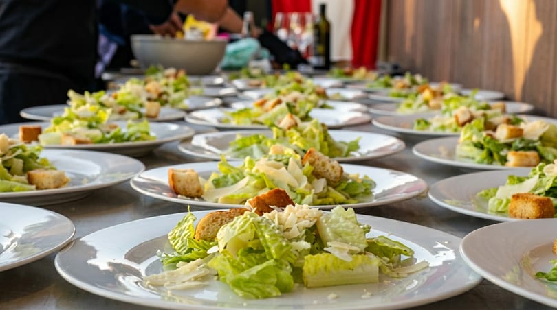 Numerous plates of fresh Caesar salad with croutons and parmesan cheese are lined up for service.