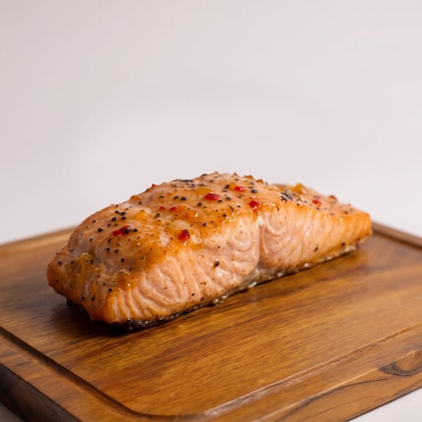 Glazed salmon fillet seasoned with pepper and red flakes on a wooden cutting board.