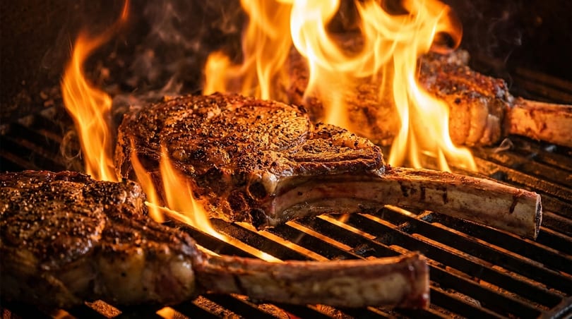 Three bone-in steaks sizzling on a grill with large, vibrant flames licking the meat.